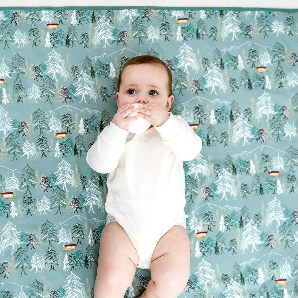 Nest Designs Cotton Waterproof Baby Change Pad Forest Fun; baby in white onesie on a teal forest-print surface.