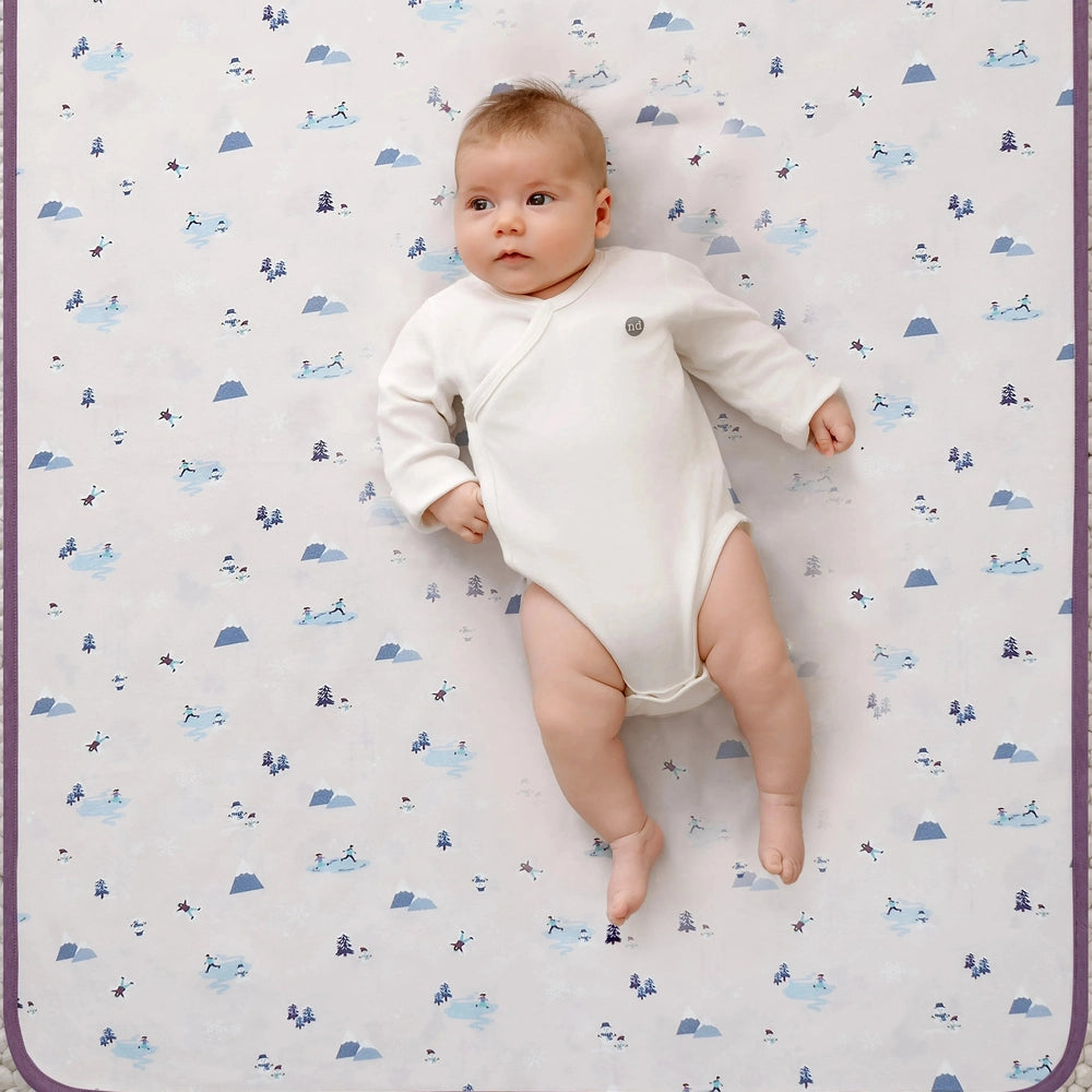 Nest Designs Cotton Waterproof Change Pad Large Snow Buddies; baby lying on white, snow-themed pad.