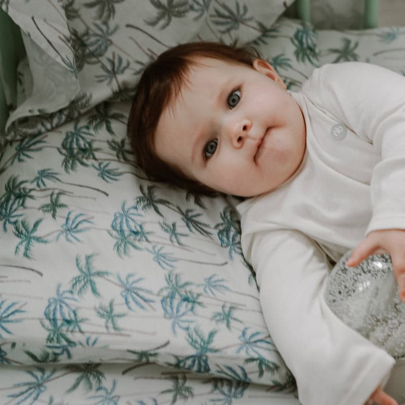 Nest Designs Foam Pillow with Bamboo Jersey Pillowcase Coco Beach; baby lying on palm-pattern bedding.