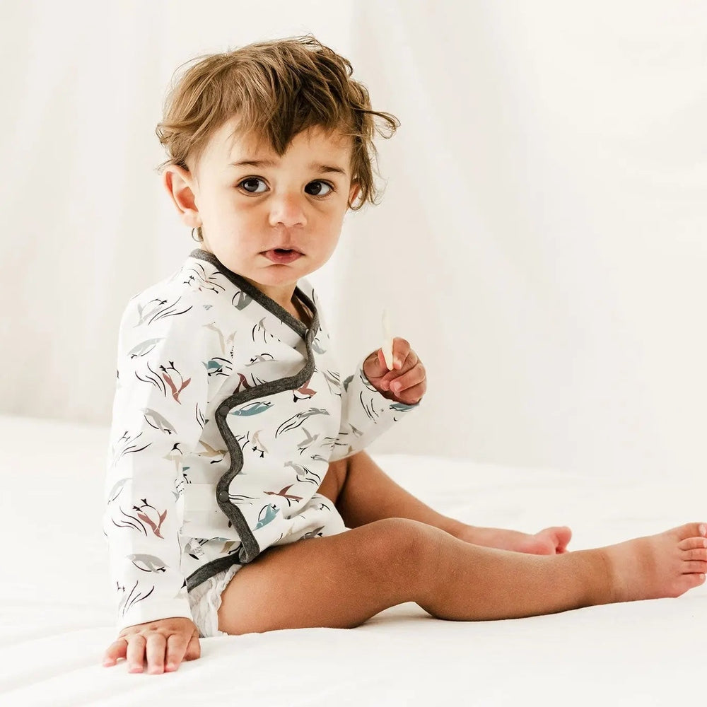 Nest Designs Organic Cotton Kimono Long-Sleeve Baby Onesie in rainbow swim print, worn by a baby sitting on a white bed.
