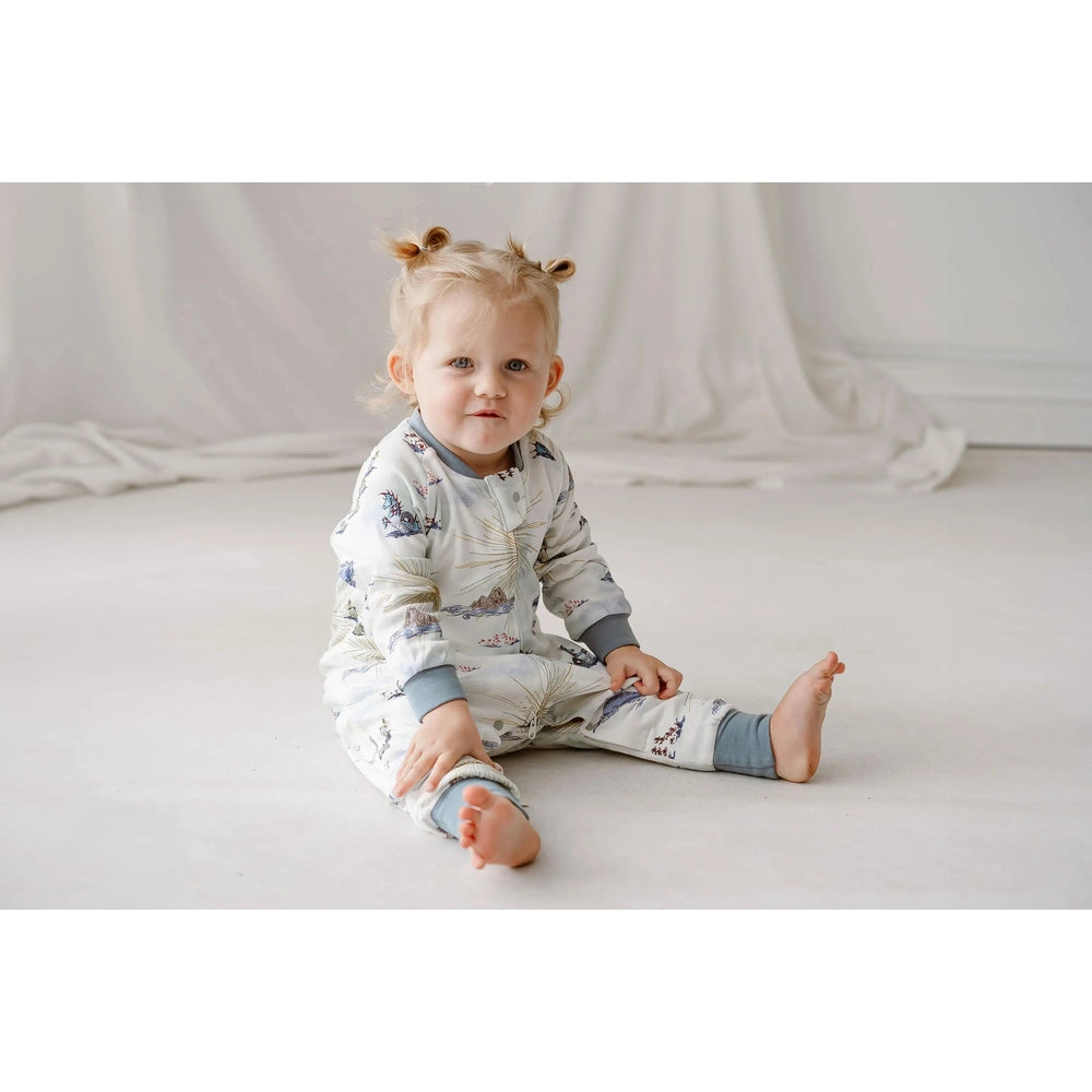 Nest Designs Organic Cotton Long Sleeve Footed Sleep Bag 1.0 TOG Ocean Wonders; baby sits on a light floor in a printed outfit