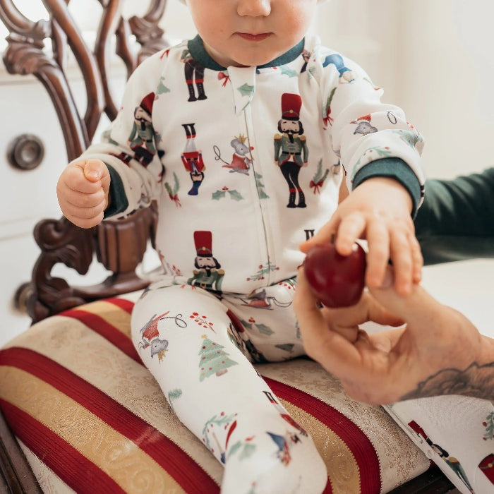 Nest Designs Organic Cotton Long Sleeve Footed Sleeper Nutcracker, white with nutcracker print; parent offers a red apple to baby.