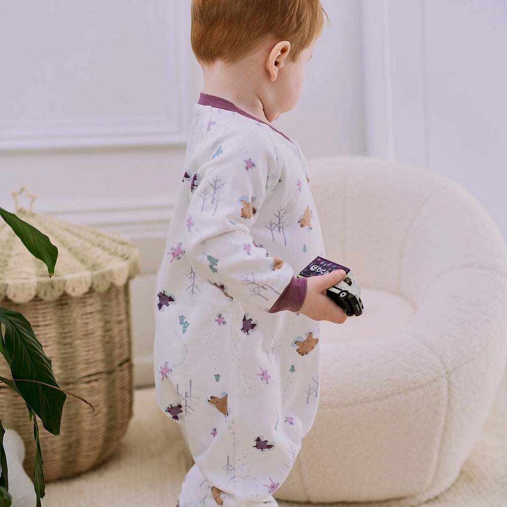 Nest Designs Organic Cotton Long Sleeve Footed Sleep Bag 1.0 Tog Snow Angels: toddler in white sleepwear holds a toy car.