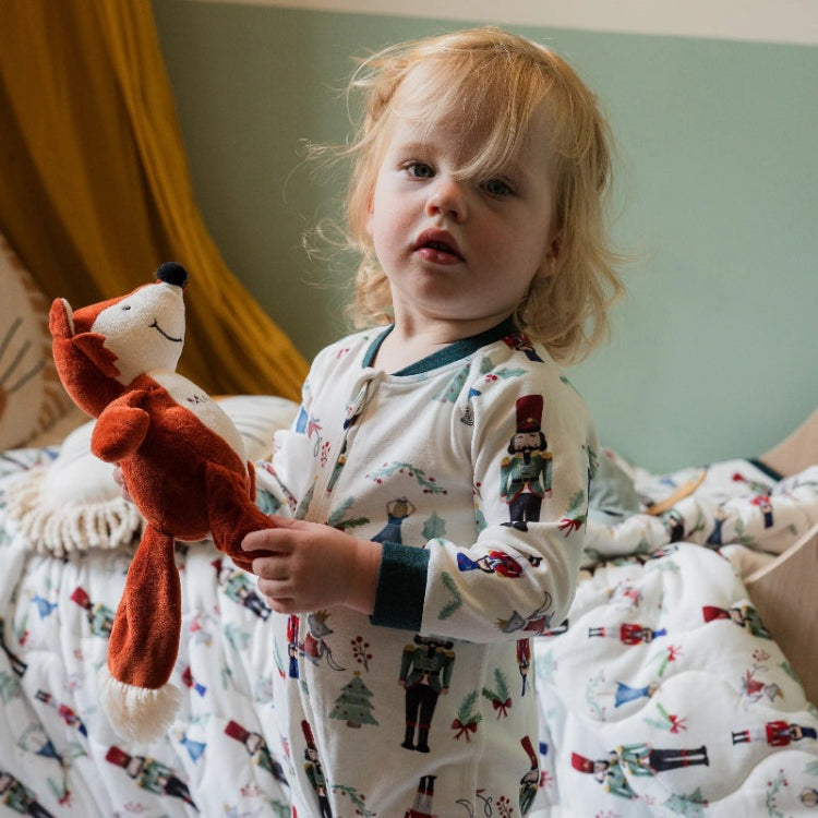 Nest Designs Organic Cotton Long Sleeve Footless Sleeper Nutcracker; toddler in white Christmas-print sleepwear holds a red plush fox.