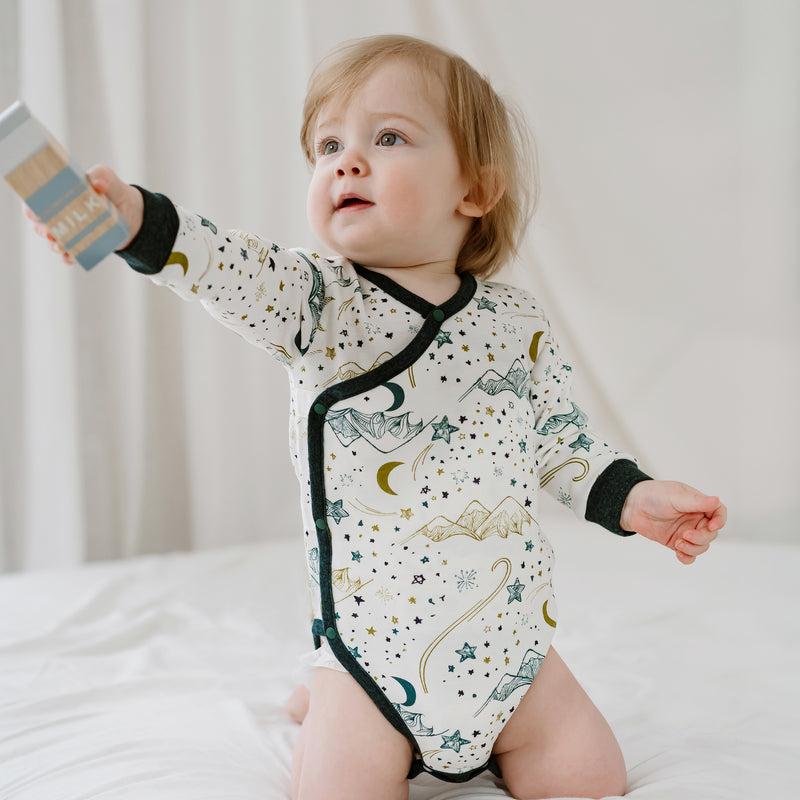 Nest Designs Organic Cotton Long Sleeve Kimono Onesie Stars White; baby on bed reaching forward, showing moon and star print.
