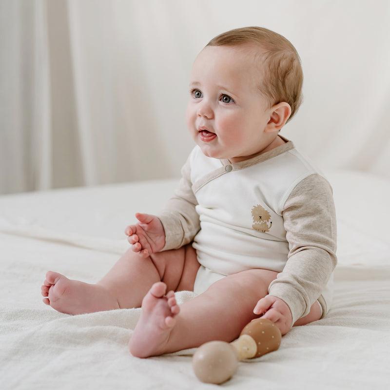 Nest Designs Organic Cotton Long Sleeve Kimono Onesie-Safawhee, cream with beige sleeves, lion logo, baby sits with wooden rattles.