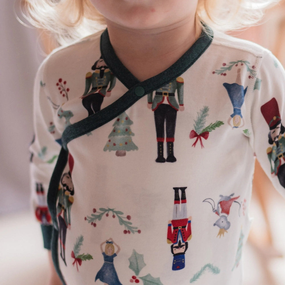 Nest Designs Organic Cotton Long Sleeve Kimono Onesie Nutcracker worn by a baby; cream fabric with Nutcracker prints and dark green wrap closure.