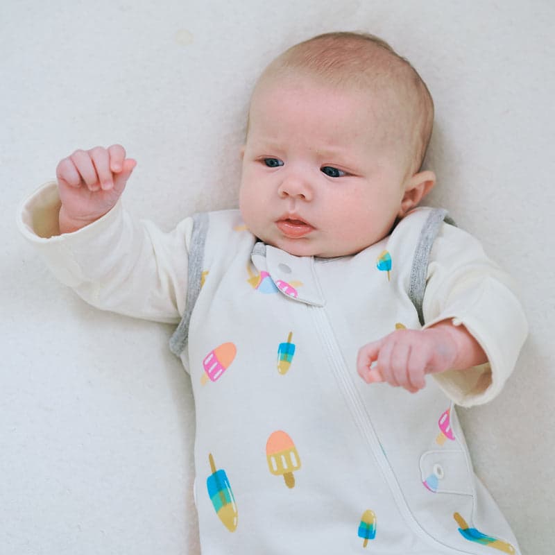 Nest Designs Startle-Stop Sleep Bag 0.5 Tog in white organic cotton with popsicle prints; baby lies on back with zipper closed.