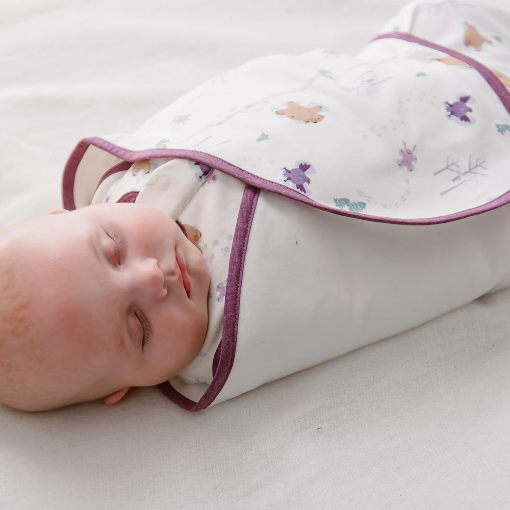 Nest Designs Organic Cotton Swaddle Sleep Bag 0.5 Tog Snow Angels; baby snugly swaddled in white fabric with purple trim.