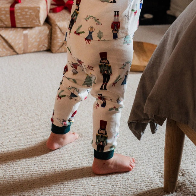 Nest Designs Organic Cotton Nutcracker two-piece long-sleeve PJ set; toddler stands barefoot on carpet in festive print.