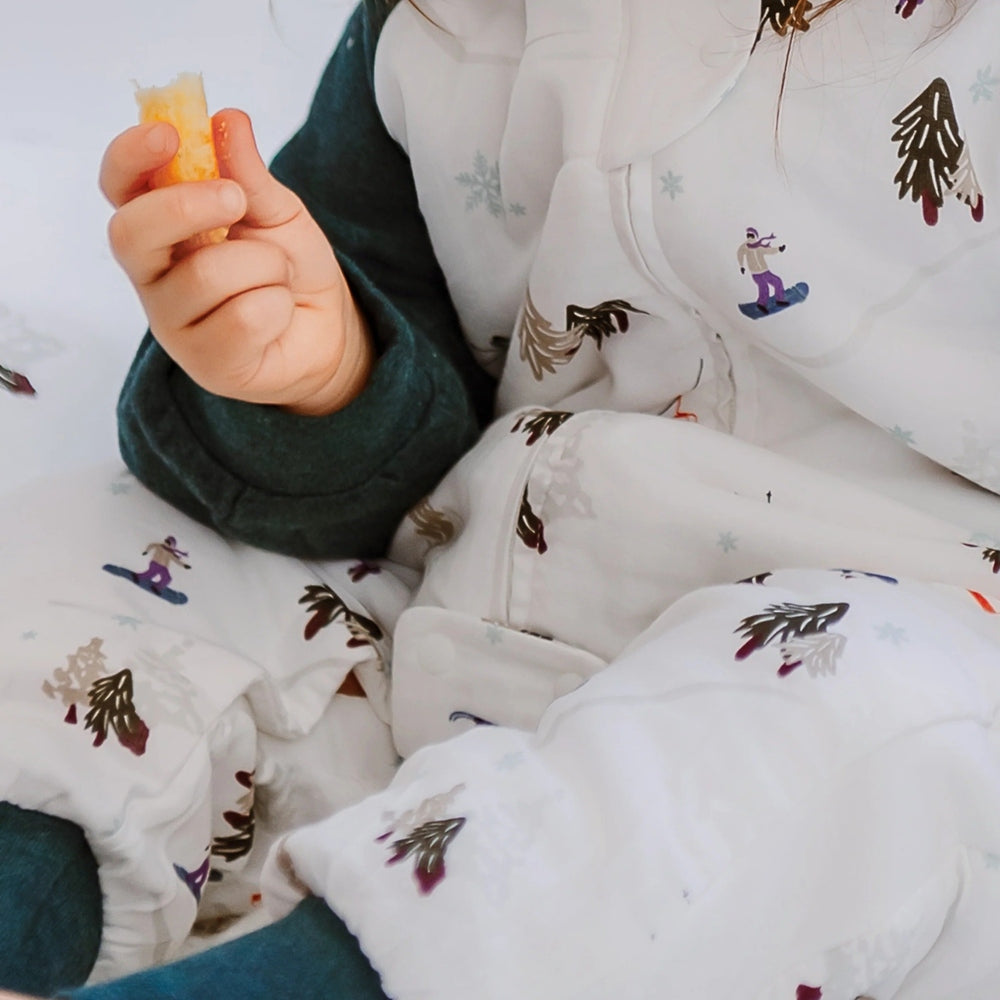 Nest Designs Raglan Bamboo Long Sleeve Footed Sleep Bag 2.5 Tog; baby in white sleep bag with winter prints, green sleeves visible, munching a snack.