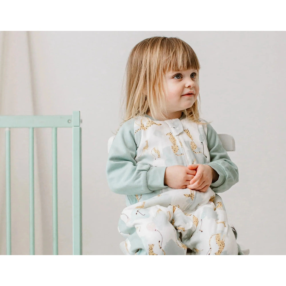Nest Designs Raglan Bamboo Long Sleeve Footed Sleep Bag 2.5 Tog Cheetah Party; toddler sits in light-blue cheetah-print sleep sack.