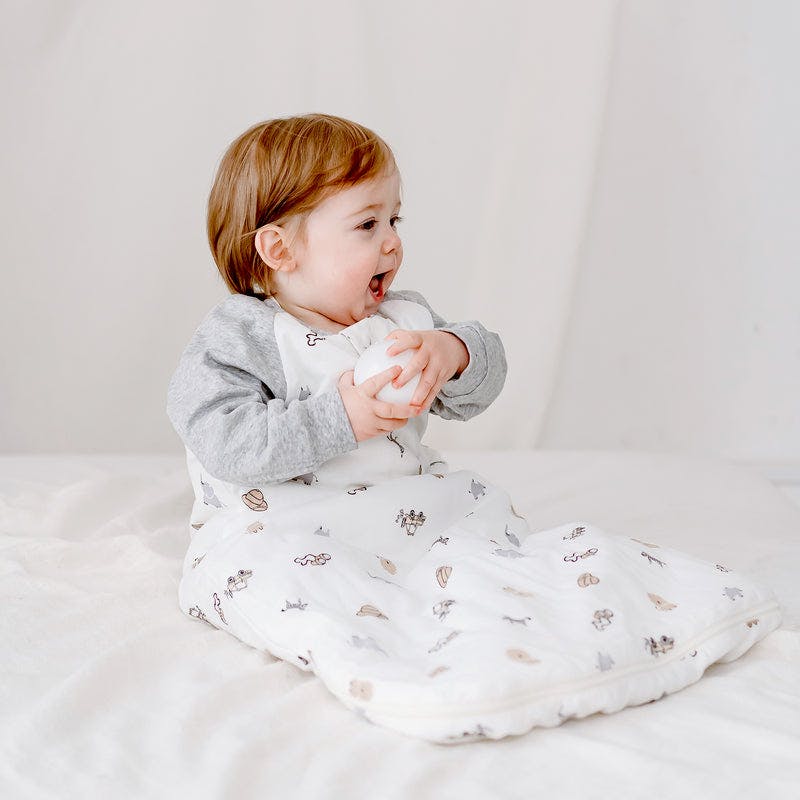 Nest Designs Raglan Bamboo Long Sleeve Sleep Bag 2.5 Tog, baby sits on a bed wearing the white sleep bag with gray sleeves, holding a ball.