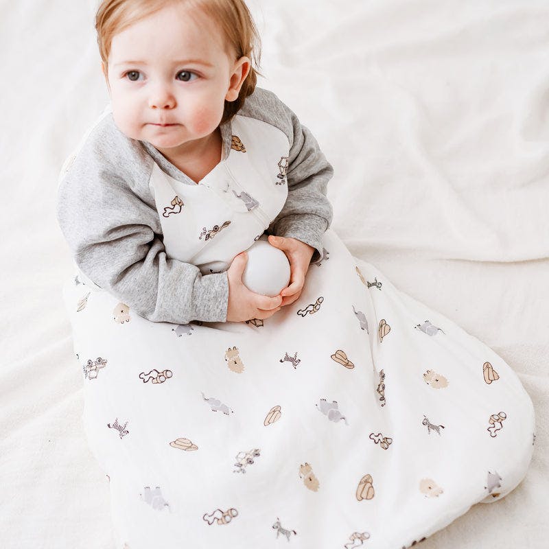 Nest Designs Raglan Bamboo Long Sleeve Sleep Bag 2.5 Tog worn by a baby cozily sitting up in a white sleep sack with gray sleeves.