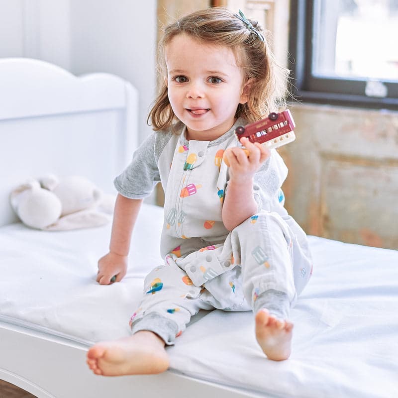 Nest Designs Raglan Bamboo Pima short-sleeve footed sleep bag in Summer Pops; toddler sits on a bed, holding a toy train.