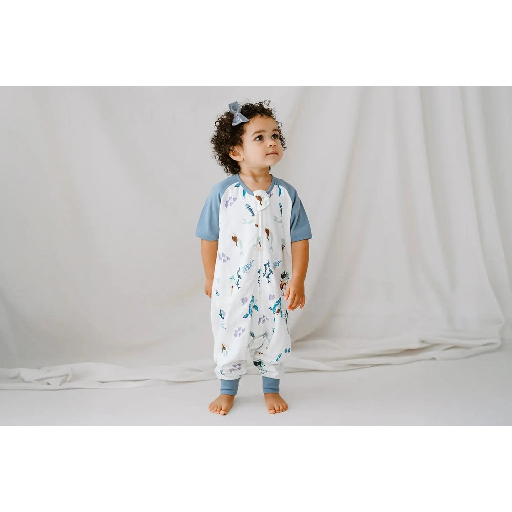 Nest Designs Raglan Bamboo Pima Short Sleeve Footed Sleep Bag 0.6 Tog Splish Splash; toddler stands in blue-sleeve white sleep bag.