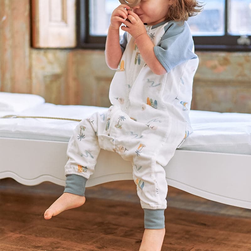 Nest Designs Raglan Bamboo Pima Short Sleeve Footed Sleep Bag 0.6 Tog Pastel Waves; white with blue cuffs, toddler calmly seated on bed.