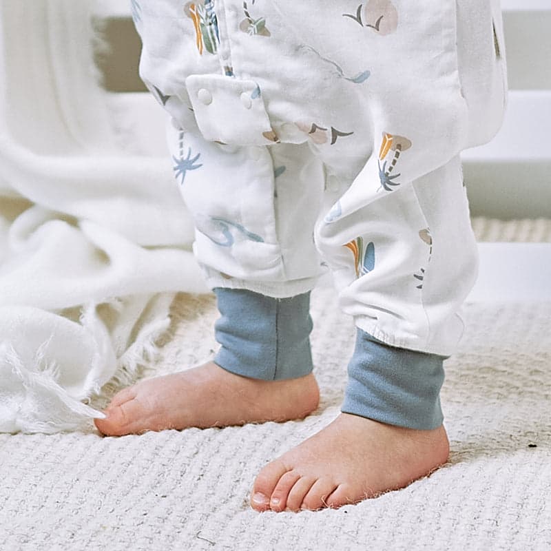 Nest Designs Raglan Bamboo Pima Short Sleeve Footed Sleep Bag 0.6 Tog in pastel waves, baby standing on rug showing the footed design