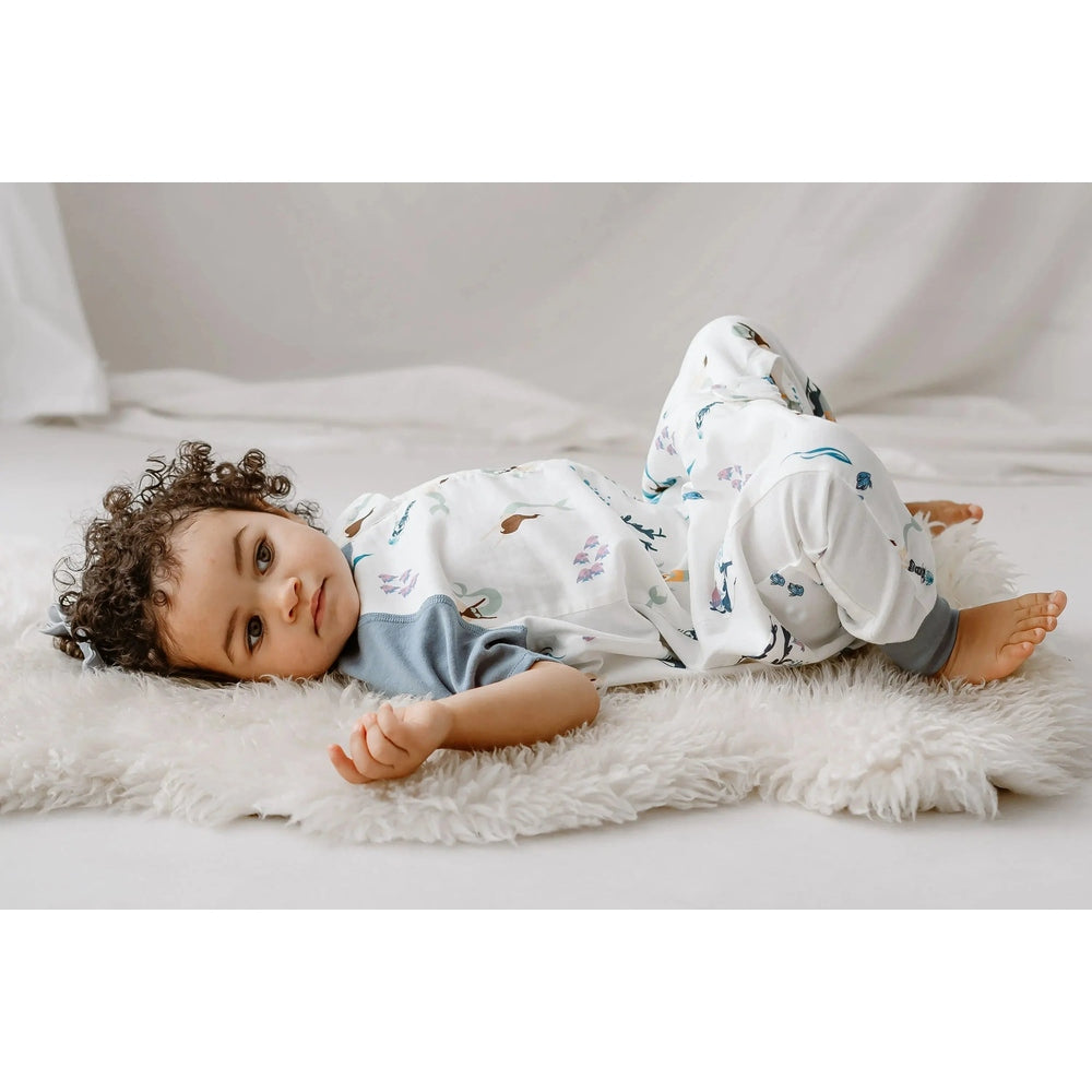 Nest Designs Raglan Bamboo Pima Short Sleeve Footed Sleep Bag 0.6 Tog Splish Splash; baby lies on a fluffy rug.