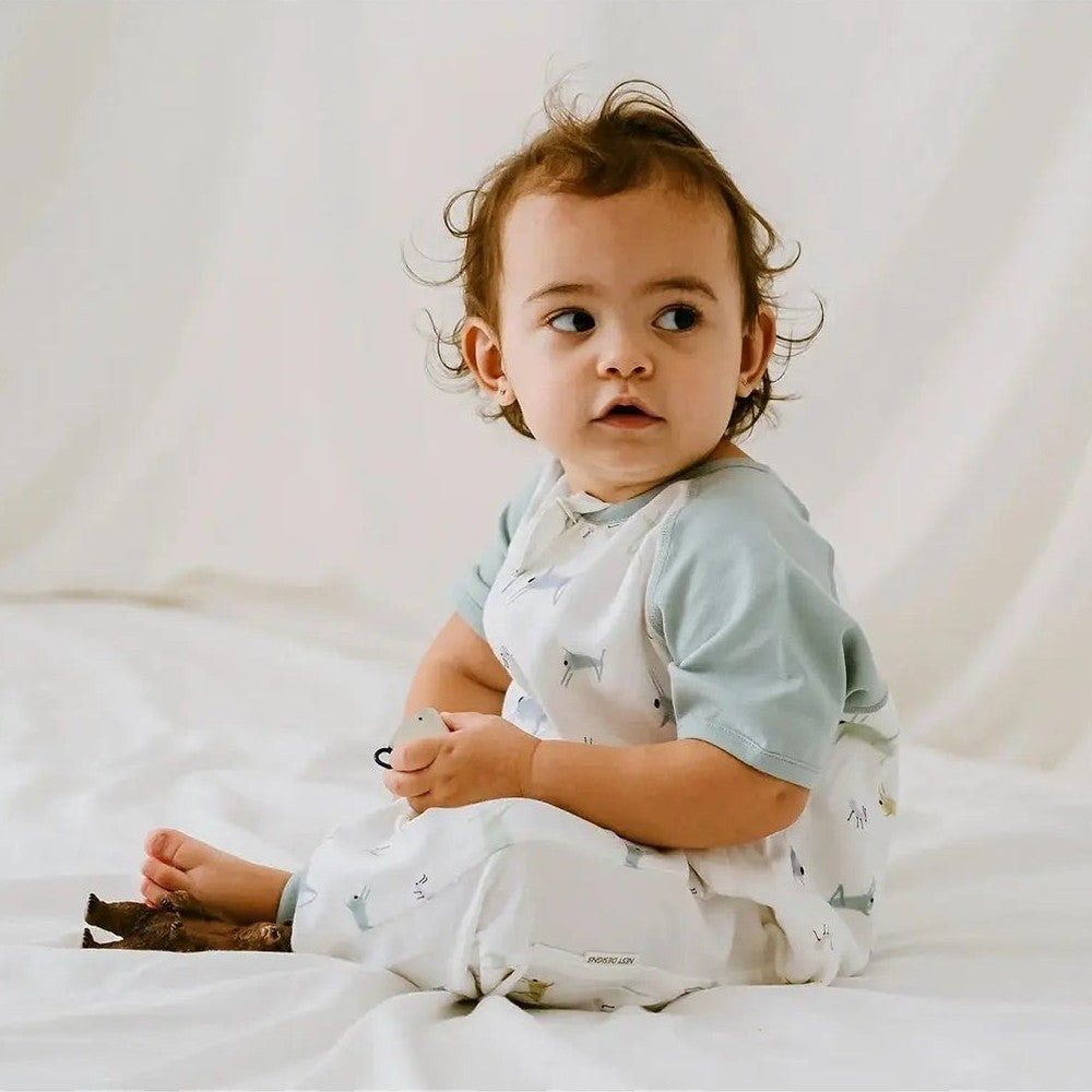 Nest Designs Raglan Bamboo Pima Short Sleeve Sleep Suit 0.6 Tog in light blue/white; baby sits on white bed.