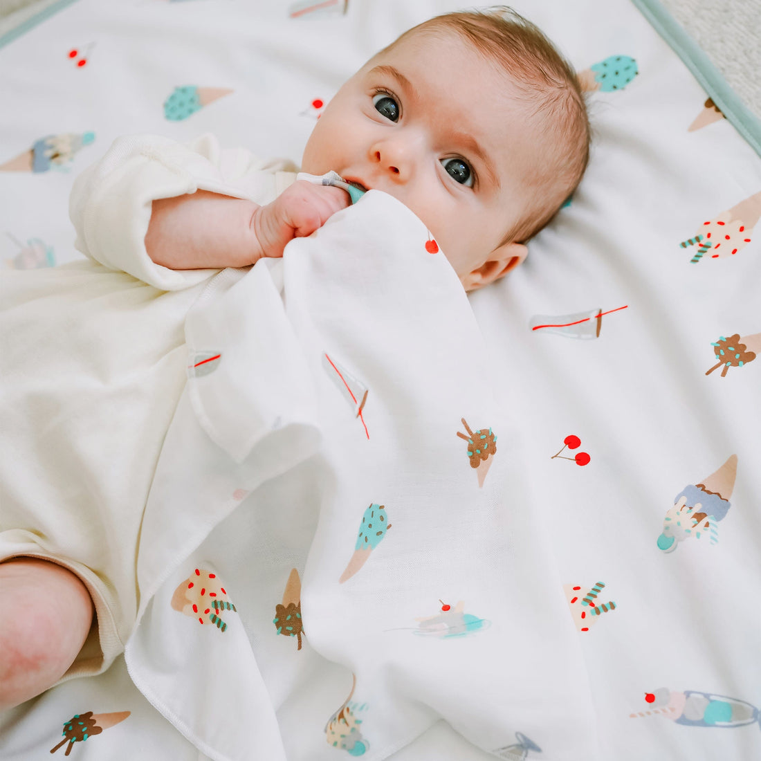 A baby lying on a white blanket with a colorful ice cream cone pattern, wearing a cream-colored onesie.