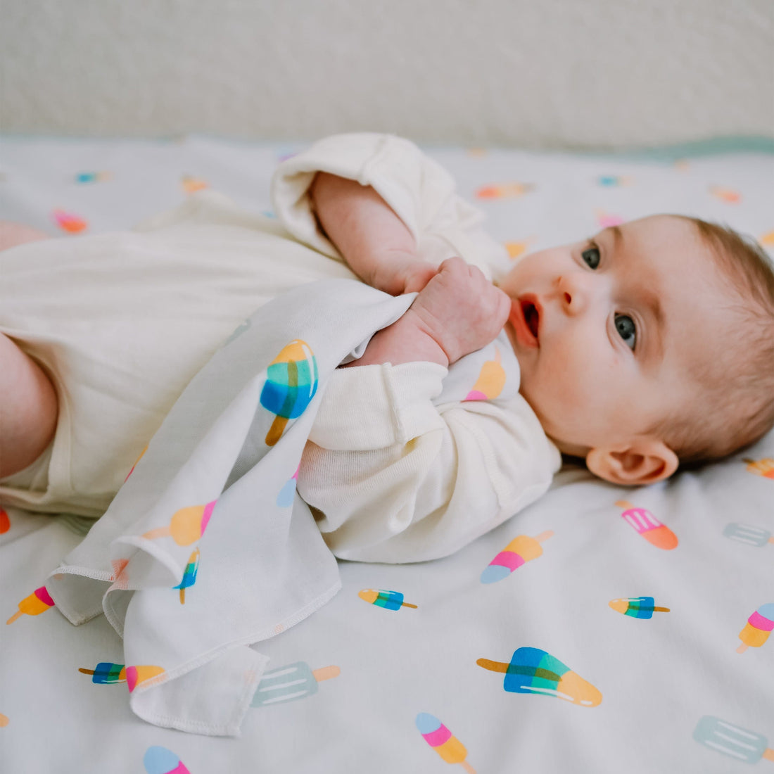 A baby lying on a white changing pad or sheet with a colorful popsicle print, wearing a cream long-sleeve bodysuit and wrapped in a matching cream cloth.