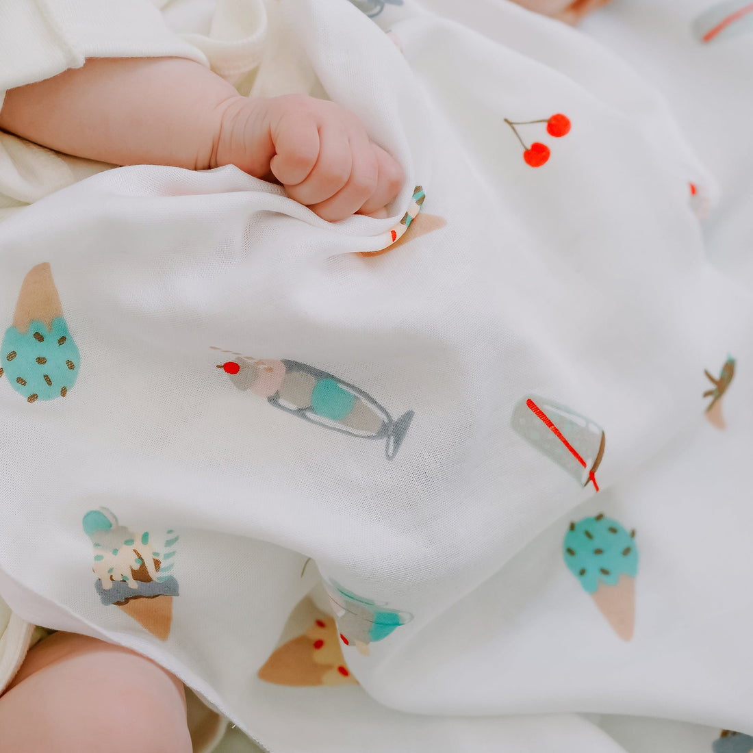 A close-up of a baby's hand and legs wrapped in a white muslin swaddle blanket printed with colorful ice cream cone and cherry illustrations.