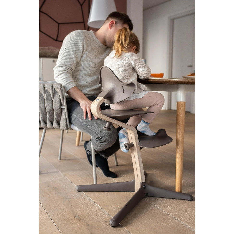 NOMI Highchair White Oak with light wood frame and gray seat, a child sits at a table while a parent assists.