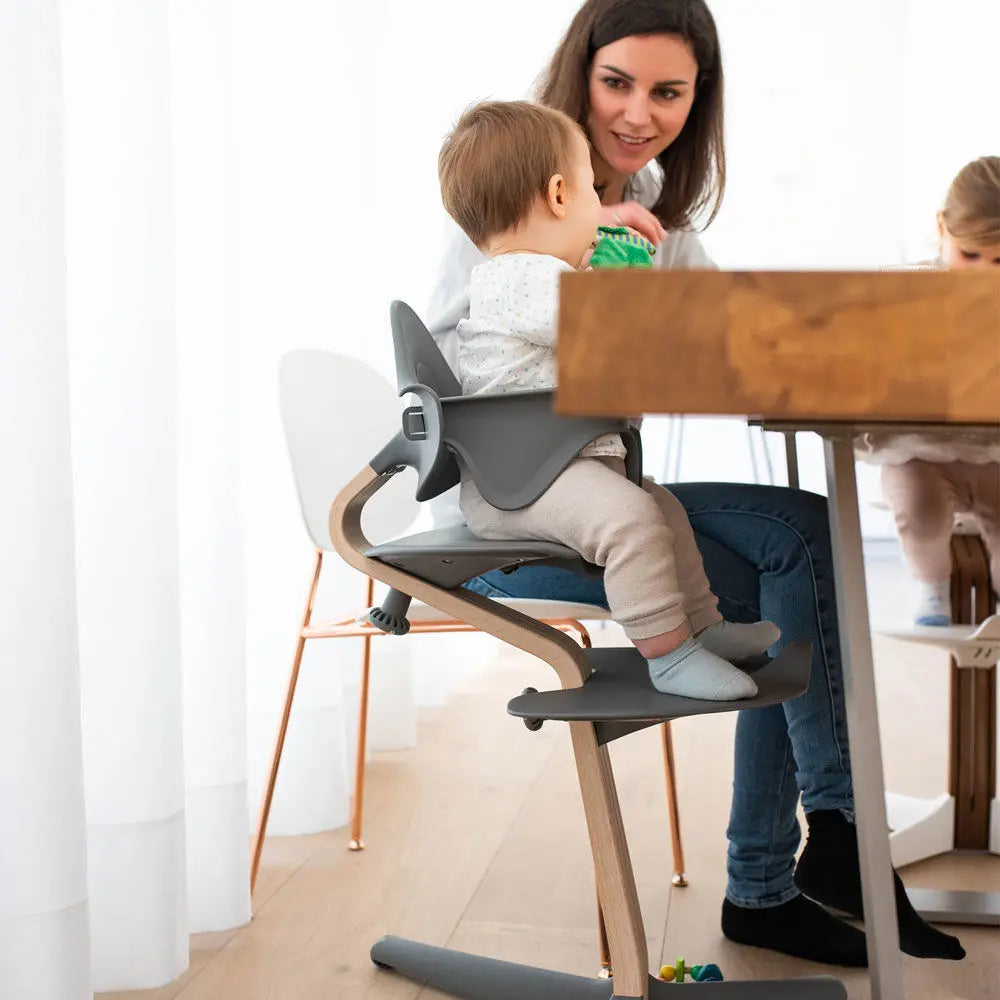NOMI Highchair White Oak with gray seat, toddler at the table as mom chats nearby.