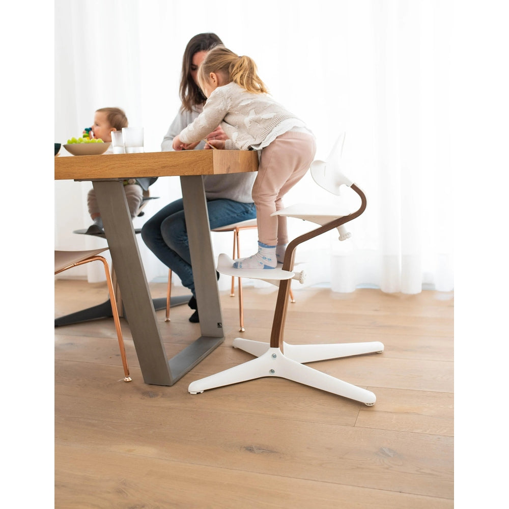 NOMI Highchair White Oak; toddler sits at the table showing wooden back/legs and white frame in use.