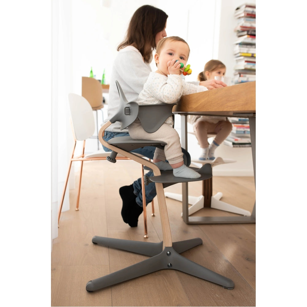 NOMI Highchair White Oak, gray seat, wooden frame; toddler sits at the dining table during mealtime.