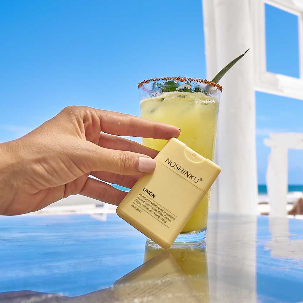 Noshinku refillable moisturizing pocket hand sanitizer beige bottle held beside a yellow cocktail on a sunny beach