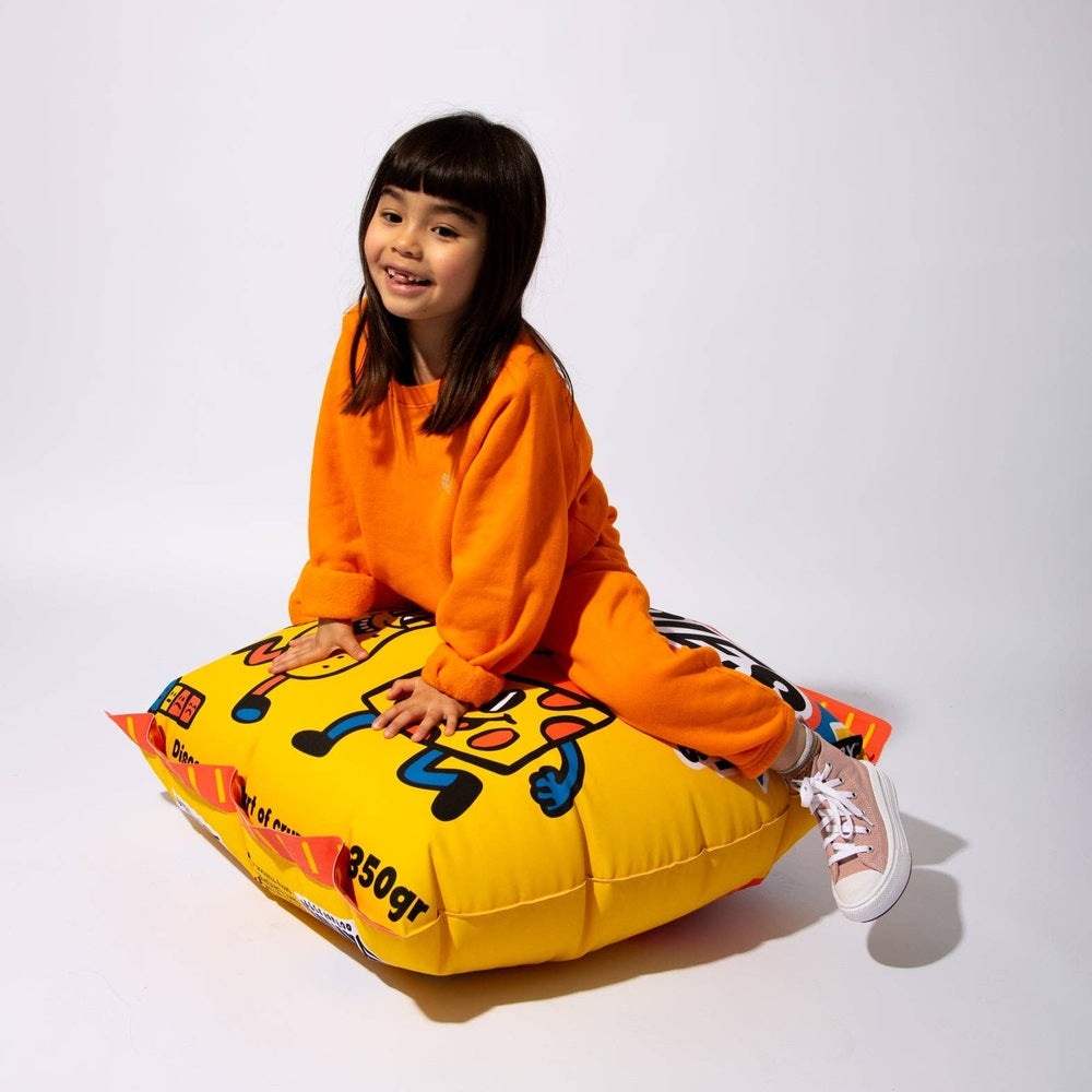OMY Crispies Giant inflatable pillow; a smiling girl in orange sits on the bright yellow cartoon-themed cushion.