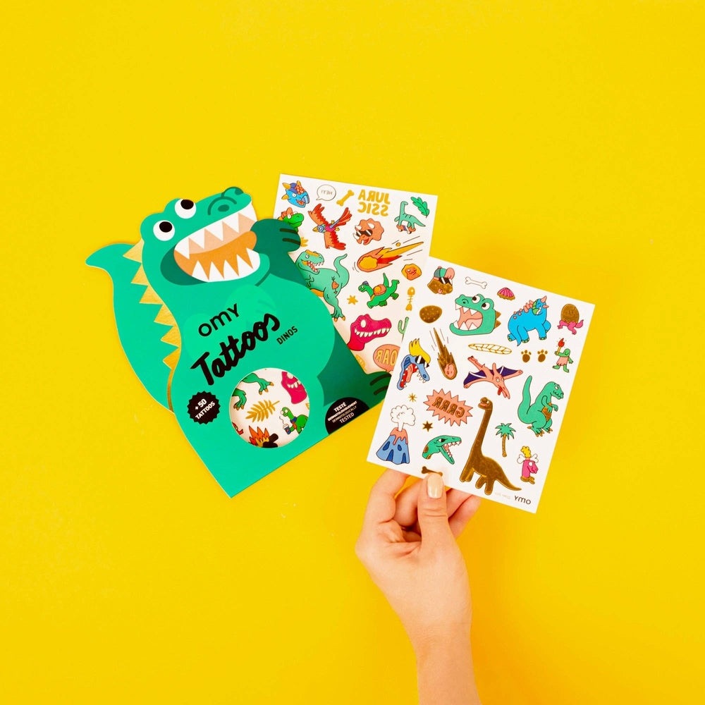 OMY Dinos Tattoos: turquoise dinosaur-shaped card with colorful temporary tattoo sheets, held by hand.