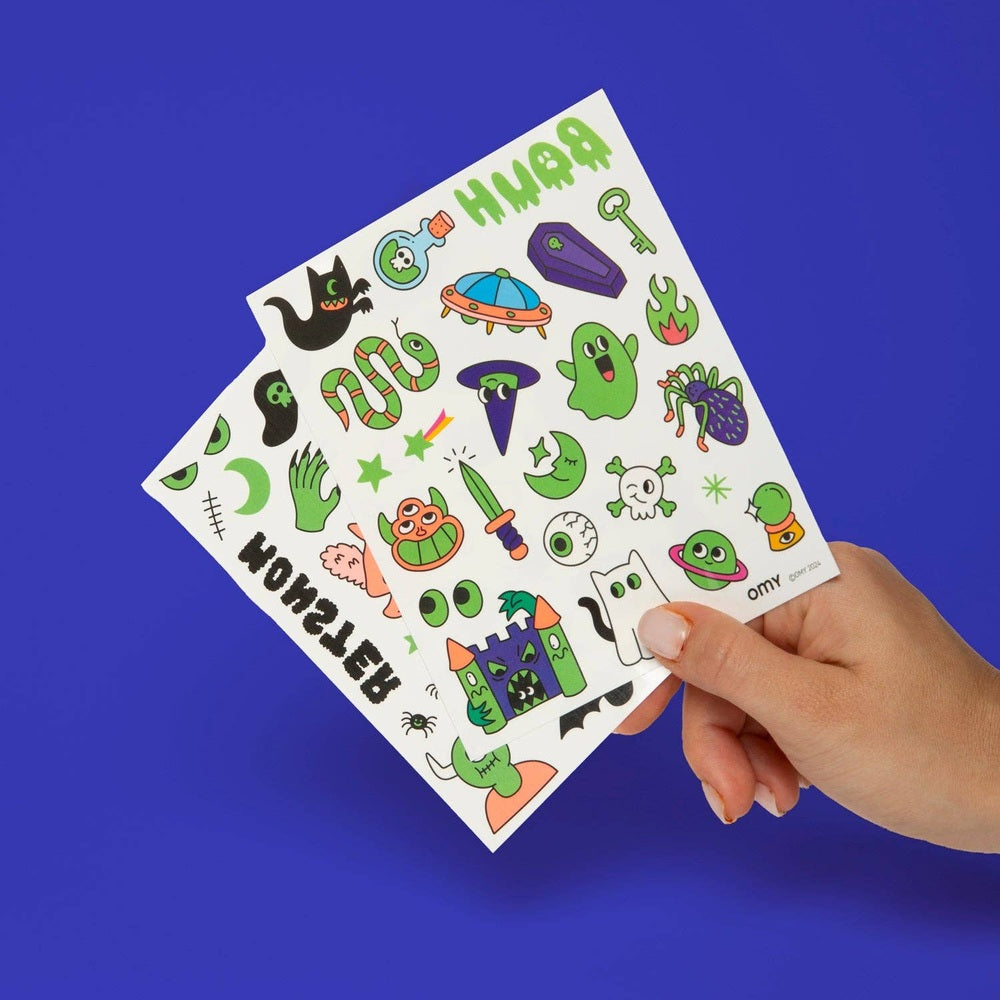 OMY Glow in the Dark Tattoos sheet with colorful monster designs, held in hand to reveal two sheets.
