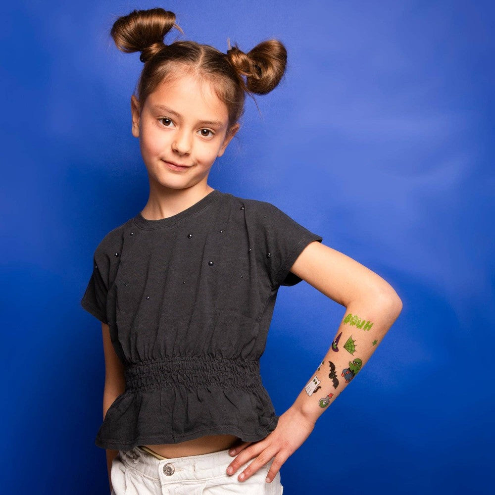 OMY Glow in the Dark Tattoos on a playful girl, showing off colorful arm tats against a blue backdrop.