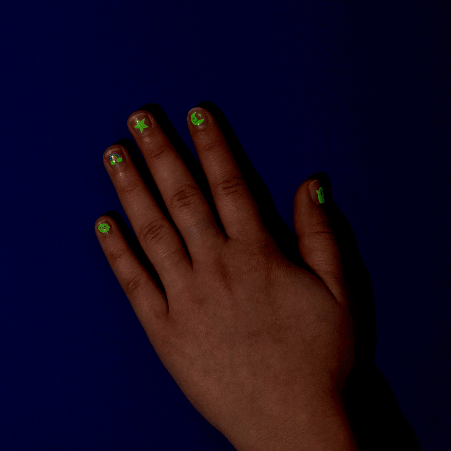 OMY Glow Party Nail Stickers on a hand with neon green star and planet designs glowing against a dark blue background.