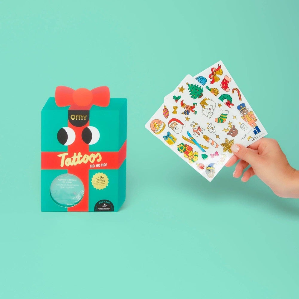 OMY Hohoho Holiday! tattoos — turquoise gift box with red bow and a hand displaying a sheet of festive temporary tattoos.