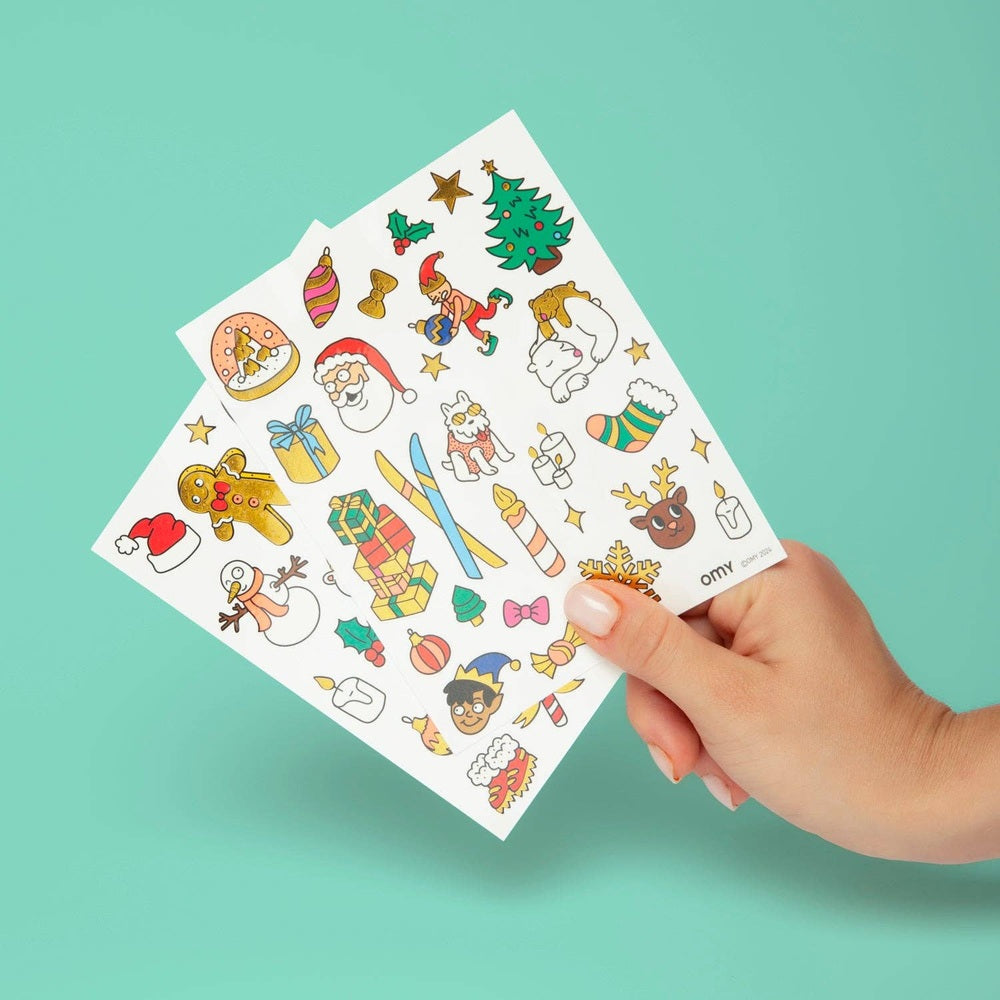 OMY Hohoho Holiday! temporary tattoos sheet held by hand featuring Santa, snowman, reindeer and gifts.