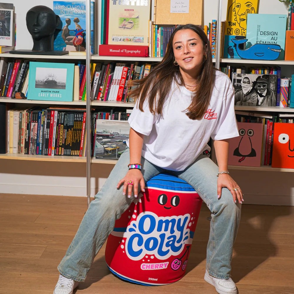 OMY Cola Giant inflatable pillow: a young woman sits on the red can-shaped cushion with blue logo among books.