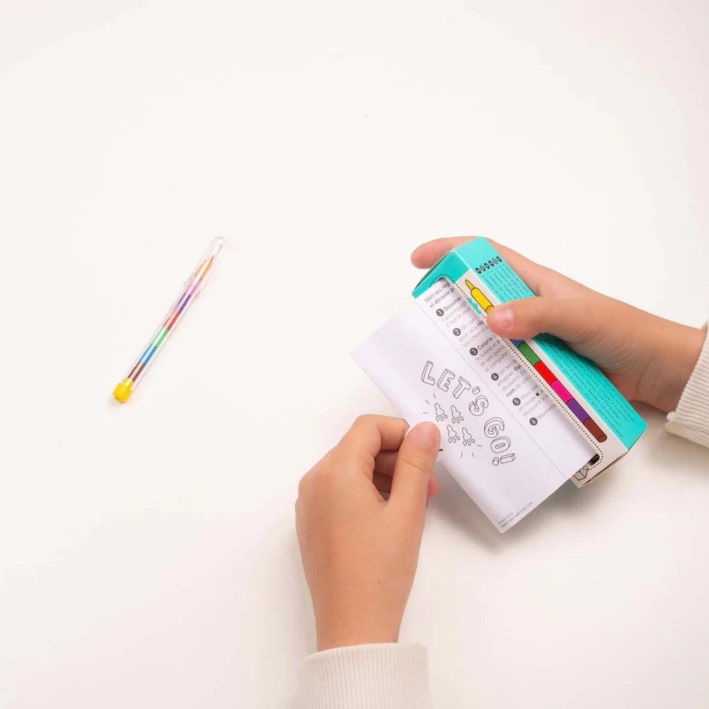OMY Pocket Game Dino coloring pad opened by hands, turquoise packaging, and rainbow-colored pencils on white desk.