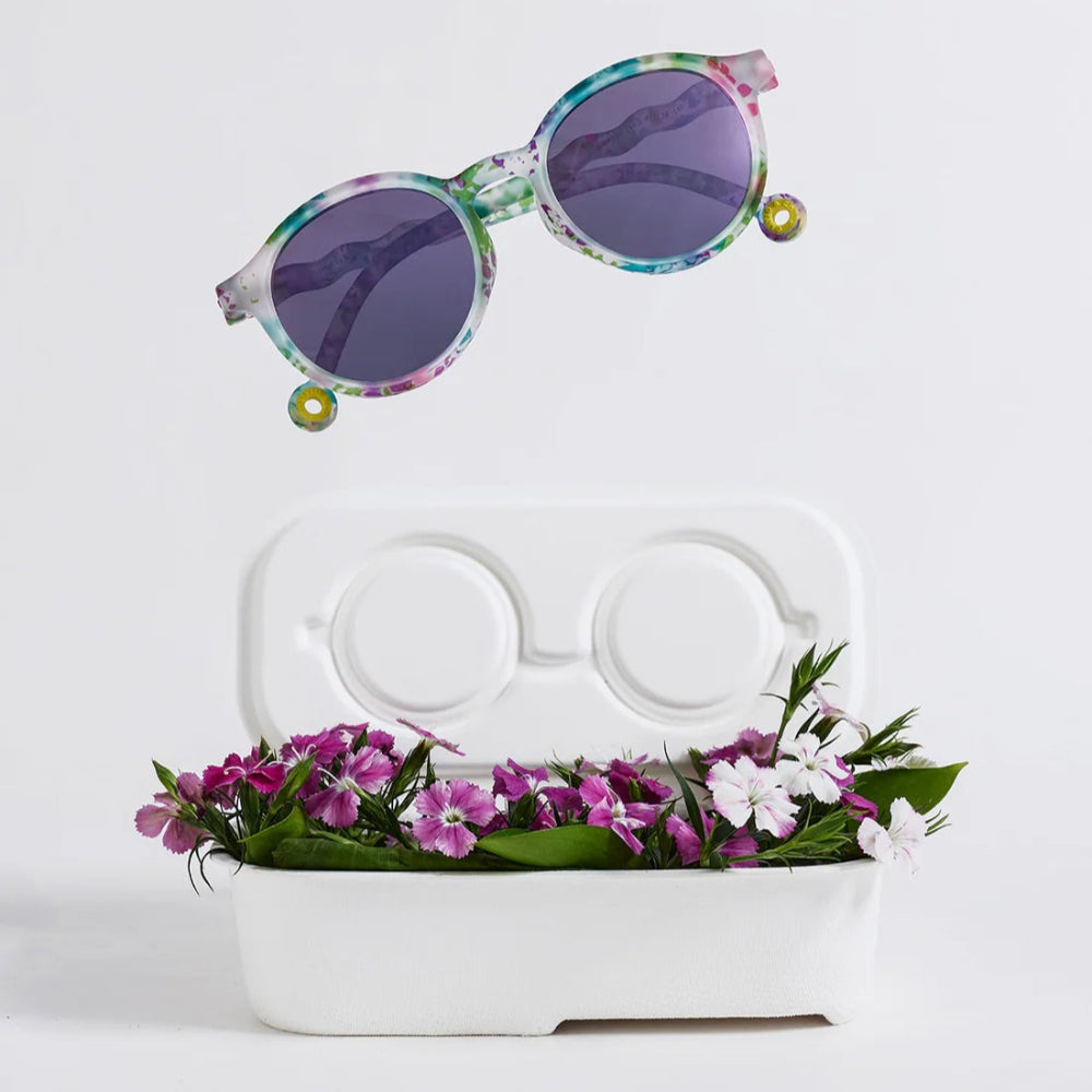 Olivio & Co Oval Sunglasses Kids Wild Flower with purple lenses, hovering above a white case beside colorful flowers.