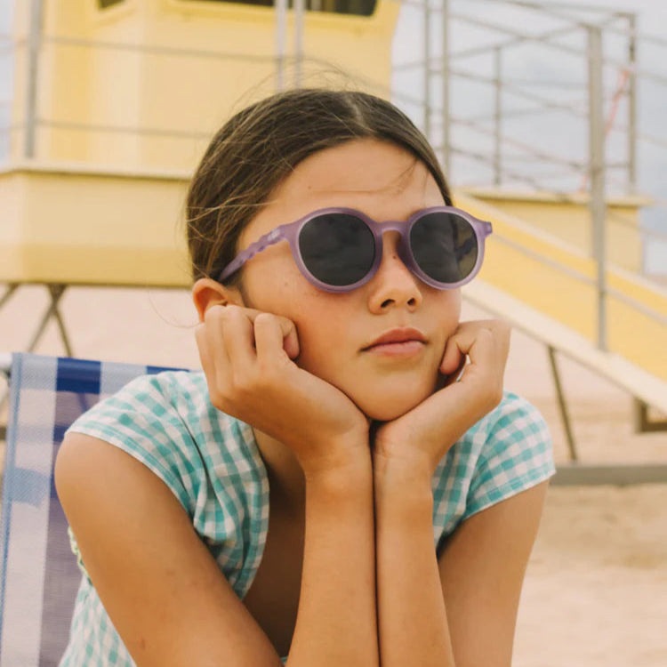 Olivio & Co Oval Sunglasses Junior Classic Olivio Mosaic Blue: girl at the beach wears purple-frame sunglasses with dark lenses.
