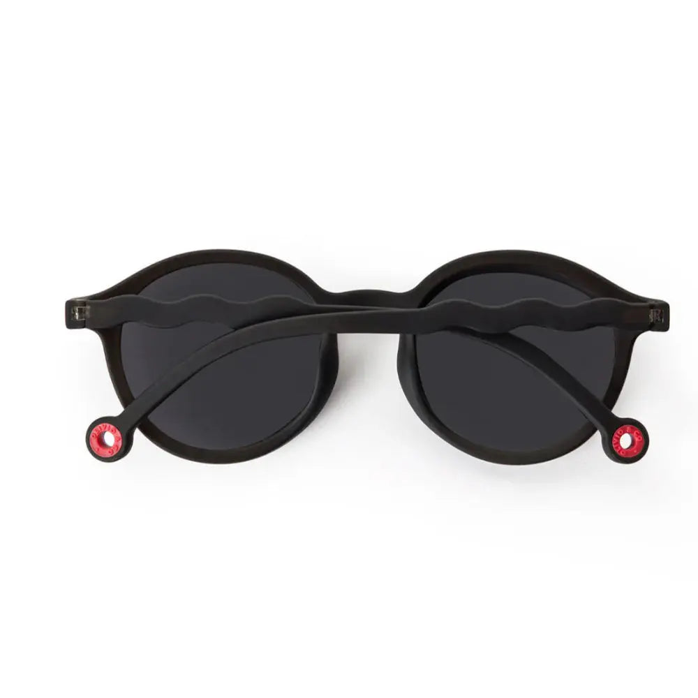 Olivio & Co Oval Sunglasses Kids/Junior Classic in black with wavy arms and red tip accents, laid on white.