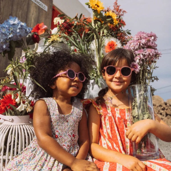Olivio & Co oval kids sunglasses with pink frames; two girls pose among a colorful flower stall.