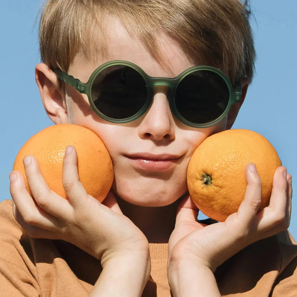Olivio & Co Round Sunglasses Junior+ Classic Olivio Olive Green; kid holds oranges to cheeks, highlighting round lenses.