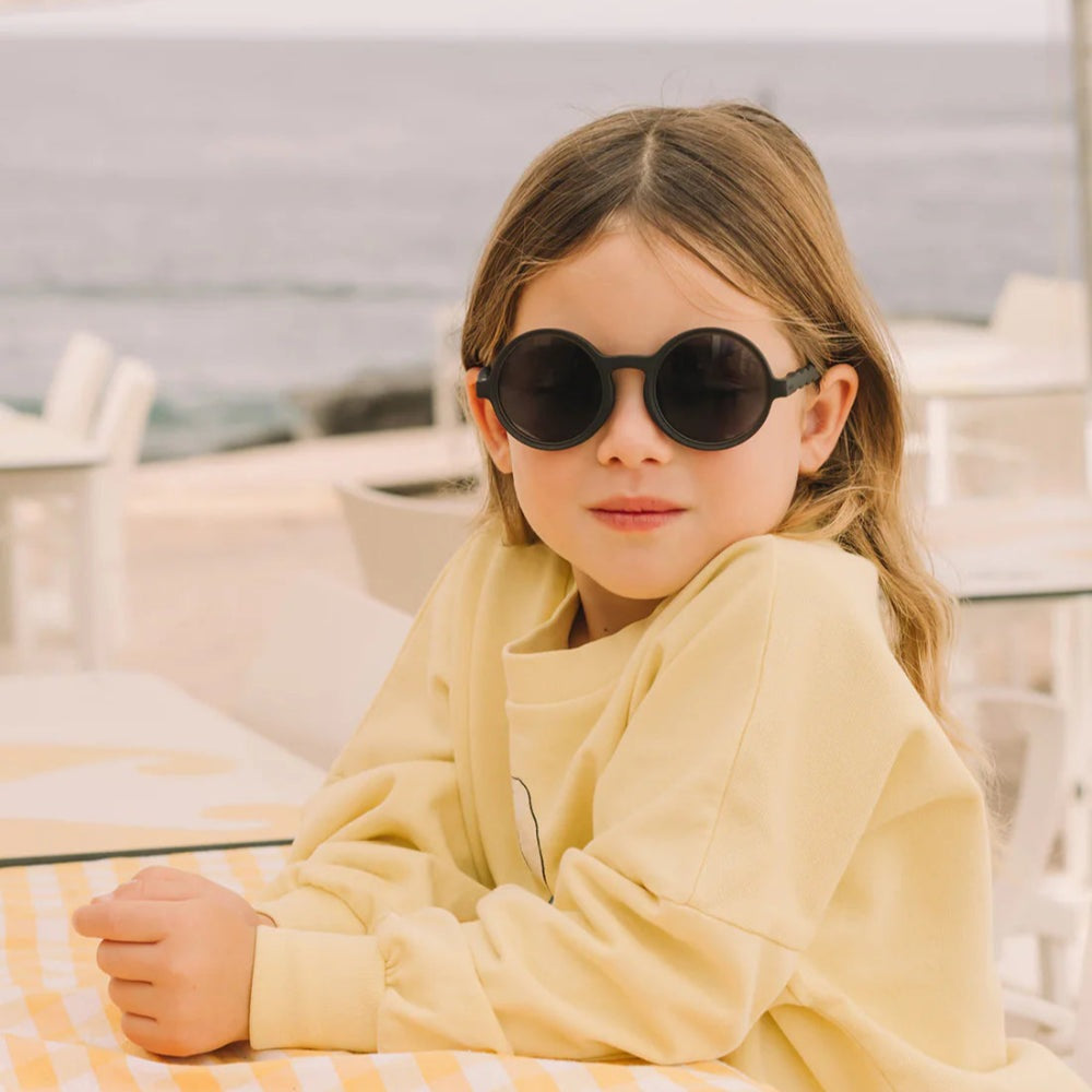 OLIVIO & CO Round Sunglasses Kids in black frame worn by a girl at an outdoor seaside cafe.