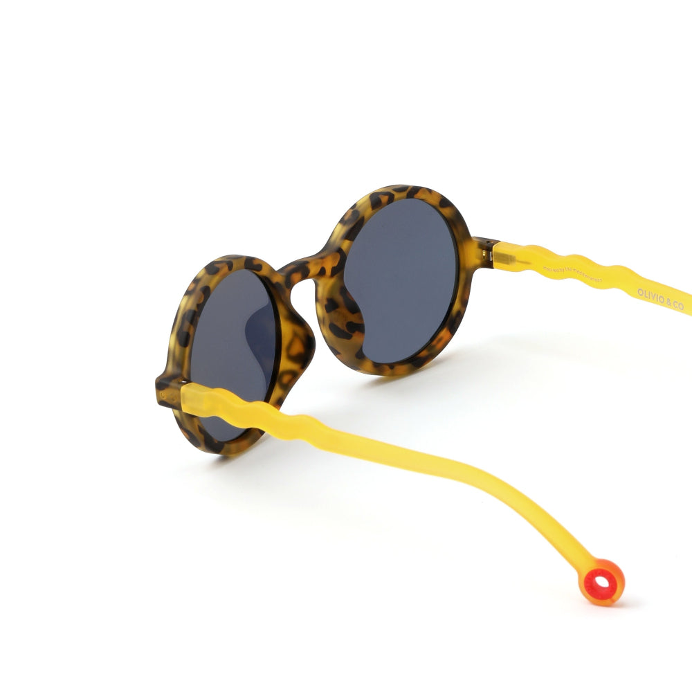 OLIVIO & CO Round Sunglasses Kids, tortoiseshell frames with bright yellow wavy arms resting on white surface