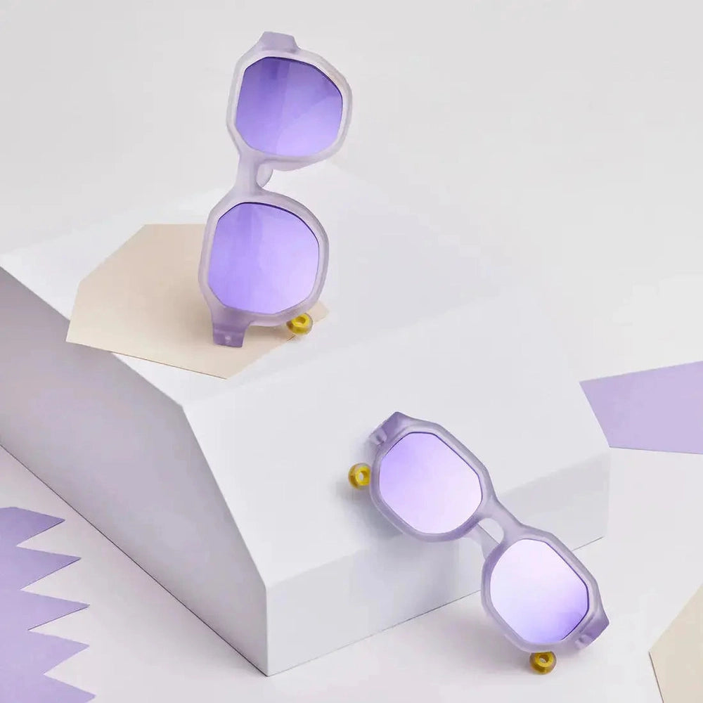 OLIVIO & CO Square Sunglasses Junior+ Creative Edition D Shell Purple; two lavender-framed sunglasses with purple lenses perched on geometric blocks.