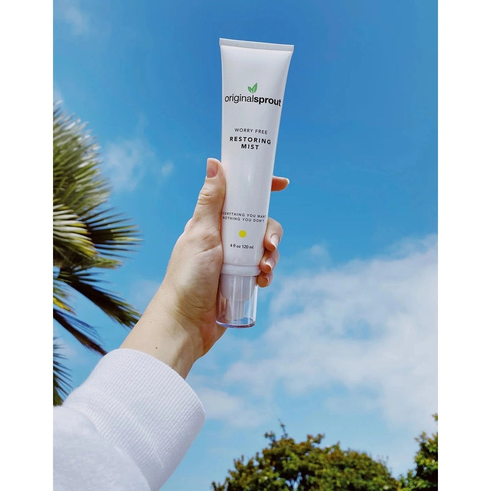 Original Sprout Worry Free Restoring Mist 4 oz white tube held in a hand outdoors against a blue sky.